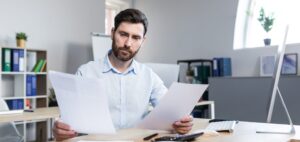 confident business man stocks broker sitting in front of monitor screen in office analyzing scaled e1676399372684